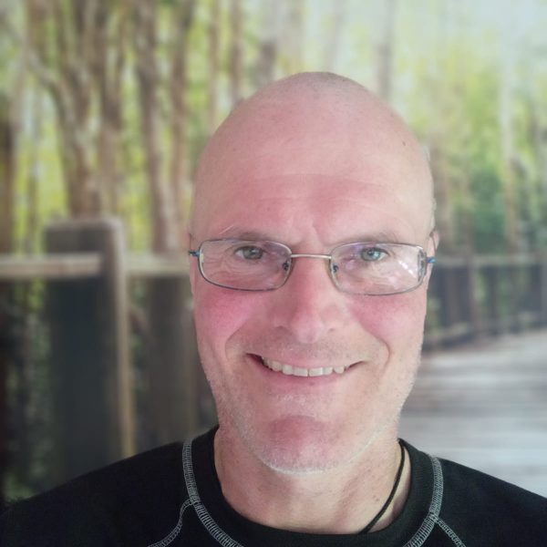 Das Bild zeigt einen kahlköpfigen Mann mittleren Alters. Der Mann trägt eine Brille und ein schwarzes T-Shirt. Das Bild ist auf Kopf-Schulter-Partie des Mannes beschränkt. Er steht vor einer Fototapete, auf der ein Steg und Bäume zu sehen sind.