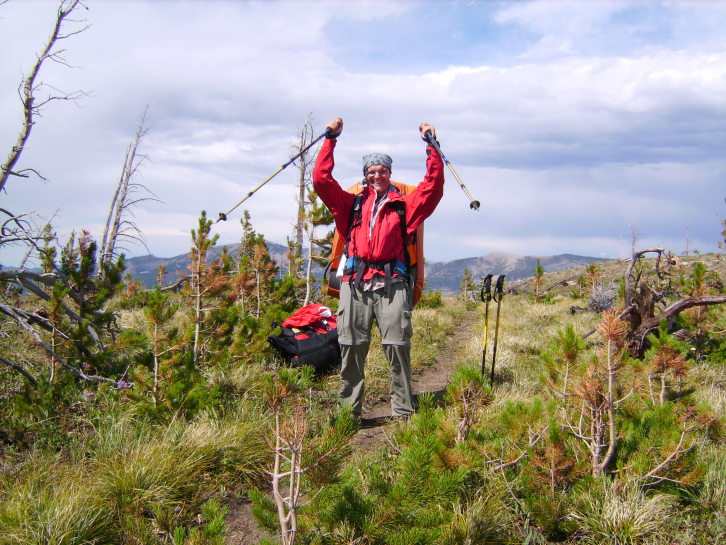 Das Bild zeigt einen Mann auf einem schmalen Wanderweg in einer wilden Landschaft. Er trägt einen großen Rucksack und hält zwei Wanderstöcke in einer Jubelpose nach oben gereckt und lacht in die Kamera.