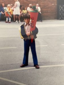 Das Bild zeigt einen Jungen bei seiner Einschulung. Er hat eine rote Schultüte im Arm, die oben mit grünem Stoff geschlossen ist. Der Junge schaut traurig in die Kamera. Das Bild wurde auf einem Schulhof aufgenommen, im Hintergrund sind weitere Menschen, darunter ein weiteres King mit einer Schultüte zu sehen.