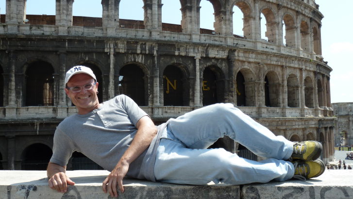 Das Bild zeigt im Vordergrund einen Mann, der seitlich auf einer Mauer liegt und in die Kamera lächelt. Im Hintergrund ist das Colosseum in Rom zu sehen, eine alte Arena.