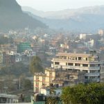 Das Bild zeigt einen Teil von Kathmandu von einer erhöhten Warte aus. Es sind viele mehrgeschossige Häuser zu sehen. Im Hintergrund sieht man Berge. Das Bild wird vom Smog im Hintergrund getrübt.