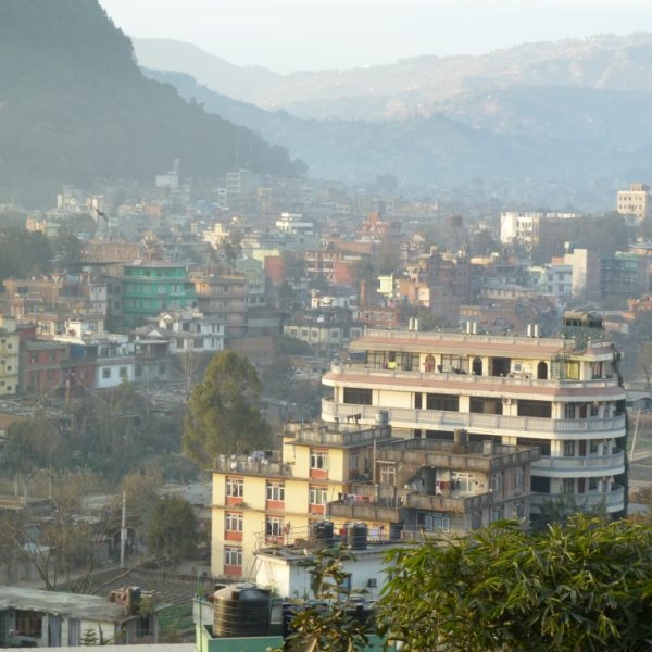 Das Bild zeigt einen Teil von Kathmandu von einer erhöhten Warte aus. Es sind viele mehrgeschossige Häuser zu sehen. Im Hintergrund sieht man Berge. Das Bild wird vom Smog im Hintergrund getrübt.