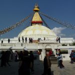 Das Bild zeigt die Stupa, ein Buddhistisches Bauwerk, welches Buddha selbst symbolisiert. Das Gebäude selbst besteht unten aus mehreren weißen Ebenen, auf denen eine gelb-weiße Kuppel aufgesetzt ist. Darauf wiederum ist ein goldener Turm, von dem Gebetsfahnen in vier Richtungen zum Boden hin gespannt sind. Vor der Stupa und auf den ersten Ebenen sind Menschen zu sehen.