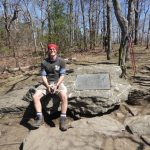 Ein Wanderer sitzt auf einem großen Stein, in den eine Plakette eingelassen ist, die den Start des Appalachian Trail anzeigt. Der Boden ist felsig, im Hintergrund sind Bäume zu sehen. An einem der Bäume lehnen zwei Paar Wanderstöcke.