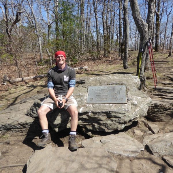 Ein Wanderer sitzt auf einem großen Stein, in den eine Plakette eingelassen ist, die den Start des Appalachian Trail anzeigt. Der Boden ist felsig, im Hintergrund sind Bäume zu sehen. An einem der Bäume lehnen zwei Paar Wanderstöcke.