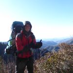 Das Bild zeigt einen warm eingepackten Wanderer mit großem Rucksack vor einer hügeligen, bewaldeten Landschaft. Der Wanderer streckt die behandschuhten Daumen in die Höhe und lächelt in die Kamera.
