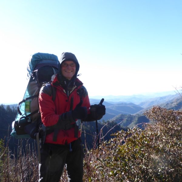 Das Bild zeigt einen warm eingepackten Wanderer mit großem Rucksack vor einer hügeligen, bewaldeten Landschaft. Der Wanderer streckt die behandschuhten Daumen in die Höhe und lächelt in die Kamera.