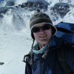 Das Bild zeigt einen Mann in warmer Kleidung, mit einer warmen Mütze und einer dunklen Sonnenbrille. Auf dem Rücken trägt er einen großen Rucksack. Im Hintergrund sind tief verschneite Berge zu sehen.