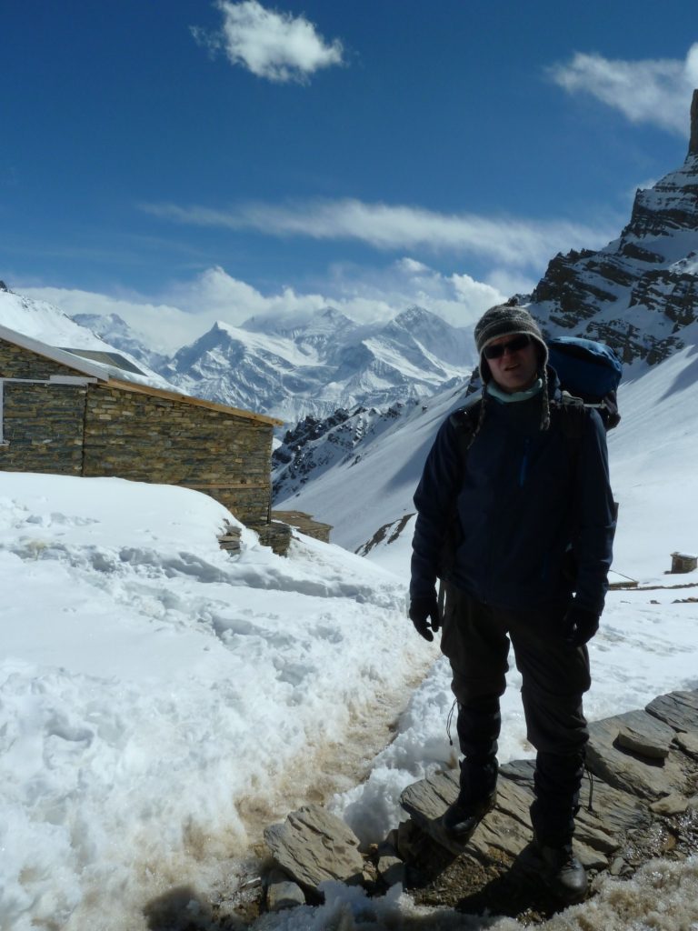 Das Bild zeigt einen sehr warm eingepackten Wanderer vor einer Bergkulisse. Alle Berge sind mit Schnee bedeckt, auf der linken Seite sieht man einen Teil einer gemauerten Hütte.