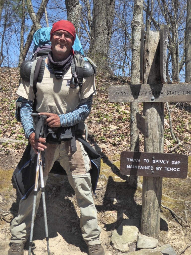 Das Bild zeigt einen Wanderer mit voller Ausrüstung und Wanderstöcken. Er steht neben einem hölzernen Wegweiser, der die Landesgrenze zwischen Tennessee und Virgina anzeigt und gleichzeitig die Entfernung zu anderen Zielen anzeigt. Im Hintergrund sind Bäume und Laub zu sehen. Auf den Schultern trägt der Wanderer Schulterpolster, um den Druck der Träger besser zu verteilen.