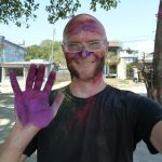 Das Bild zeigt einen Mann beim Holi-Festival in Nepal. Sein Gesicht und sein T-Shirt sind bereits leicht mit Farben bedeckt. Er lächelt in die Kamera und seine rechte Hand, mit der er in die Kamera winkt, ist lila gefärbt.