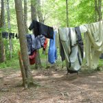 Das Bild zeigt eine kleine Lichtung in einem Wald. Zwischen Bäumen ist eine Leine gespannt. Auf dieser Leine hängen verschiedene Gegenstände, wie zum Beispiel en Zelt, ein Schlafsack, Wanderschuhe und Kleidung, zum austrocknen.