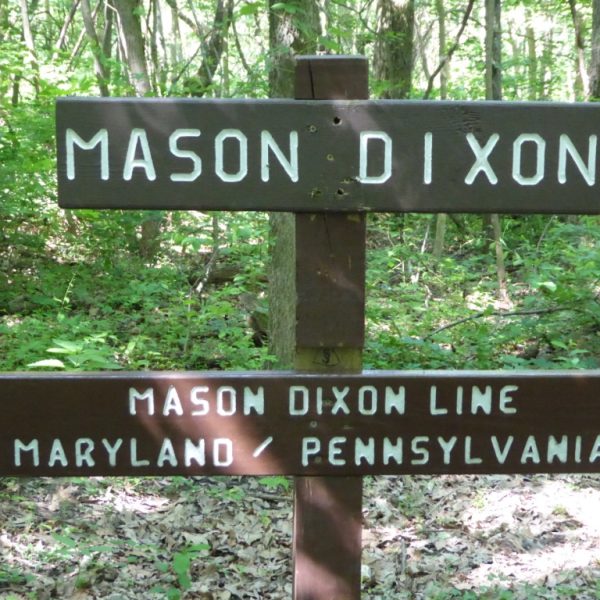 Das Bild zeigt im Vordergrund in Großaufnahme einen hölzernen Wegweiser. Auf ihm sind übereinander zwei Schilder angebracht. Das obere ist mit "Mason Dixon" beschriftet, auf dem unteren steht "Mason Dixon Line", darunter "Maryland / Pennsylvania". Im Hintergrund sind Bäume zu sehen.