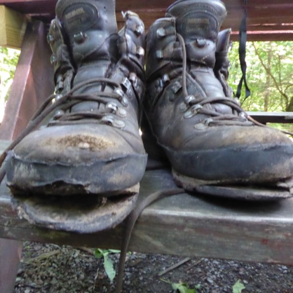 Das Bild zeigt ein paar Wanderschuhe auf einer Holzbank in Großaufnahme. Die Schuhe sind im vorderen Teil kaputt, die Sohlen haben sich schon sehr von den Schuhen gelöst. Im Hintergrund ist Wald zu erkennen.