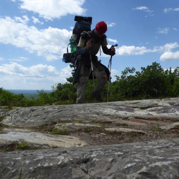 Das Bild zeigt einen Wanderer in Bewegung, wie er mit vollem Rucksack ein Stück Fels heraufläuft. Im Hintergrund ist blauer Himmel mit ein paar weißen Wolken zu sehen. In der Mitte des Bildes sind Bäume und Sträucher zu erkennen.