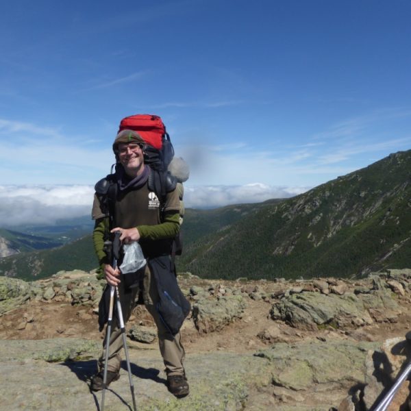 Das Bild zeigt einen Wanderer vor einer Bergkulisse. Die Hälfte des Bildes nimmt blauer Himmel ein, im unteren Teil sind Wolken zu sehen. Der Wanderer hat einen großen Rucksack auf den Schultern und Wanderstöcke in der Hand. Im unteren rechten Teil des Bildes liegen weitere Wanderstöcke, darüber sind in einiger Entfernung Menschen zu sehen.