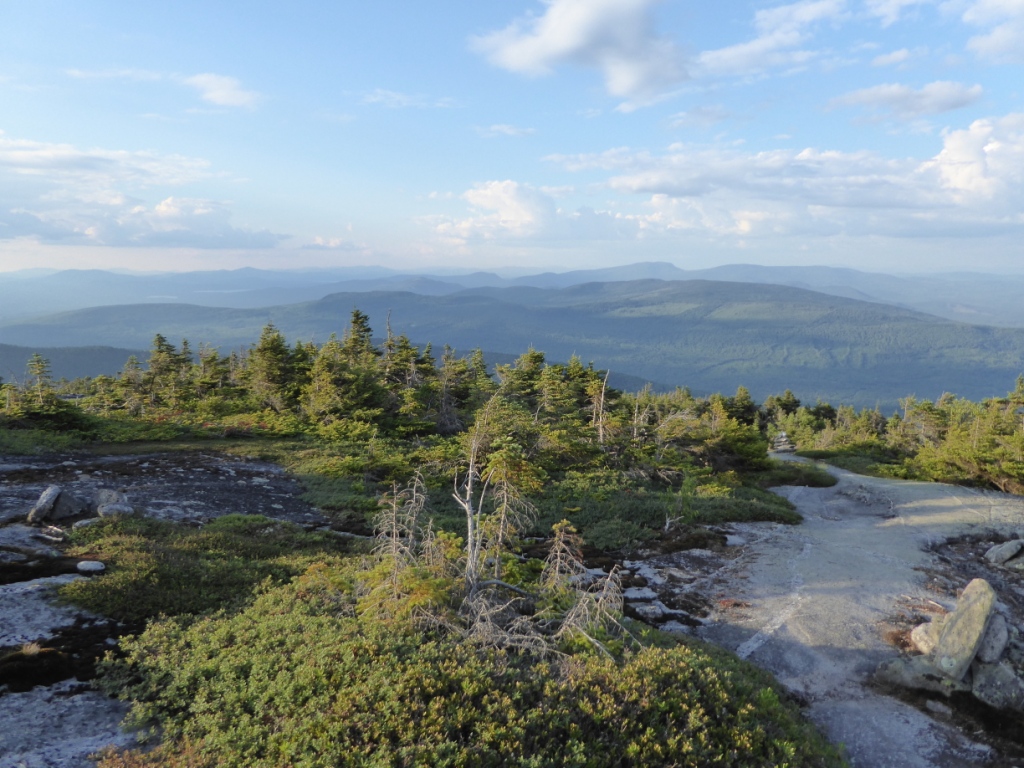 Das Bild wurde auf einem Berg aufgenommen. Es zeigt einen felsigen Weg und Sträucher. Im Hintergrund sind weitere bewaldete Hügel zu erkennen. Der Himmel nimmt die Hälfte des Bildes ein, er ist blau, mit einigen weißen Wolken durchzogen.