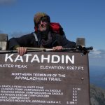 Ein Wanderer lehnt über einem großen Holzschild. Auf dem Schild ist der Gipfel des Katahdin-Berges in Maine angezeigt, die Höhe des Berges. Außerdem steht dort, dass dieser Punkt das nördliche Ende bzw. den nördlichen Start des Appalachian Trails ist. Darunter sind noch einige Kilometerangaben zu weiteren Zielen angegeben. Der Wanderer wirkt müde, er trägt einen großen Rucksack auf dem Rücken.