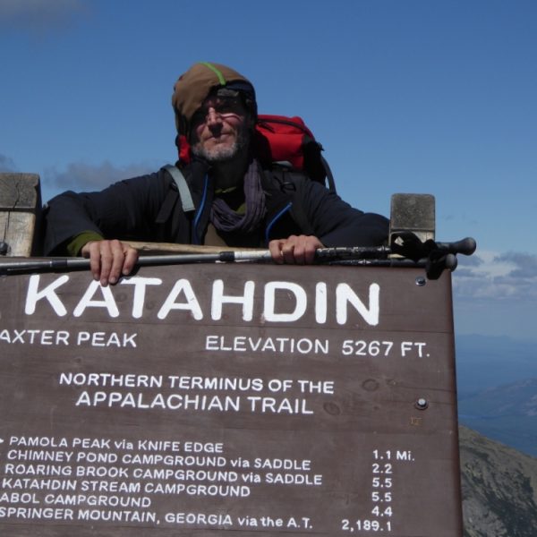 Ein Wanderer lehnt über einem großen Holzschild. Auf dem Schild ist der Gipfel des Katahdin-Berges in Maine angezeigt, die Höhe des Berges. Außerdem steht dort, dass dieser Punkt das nördliche Ende bzw. den nördlichen Start des Appalachian Trails ist. Darunter sind noch einige Kilometerangaben zu weiteren Zielen angegeben. Der Wanderer wirkt müde, er trägt einen großen Rucksack auf dem Rücken.
