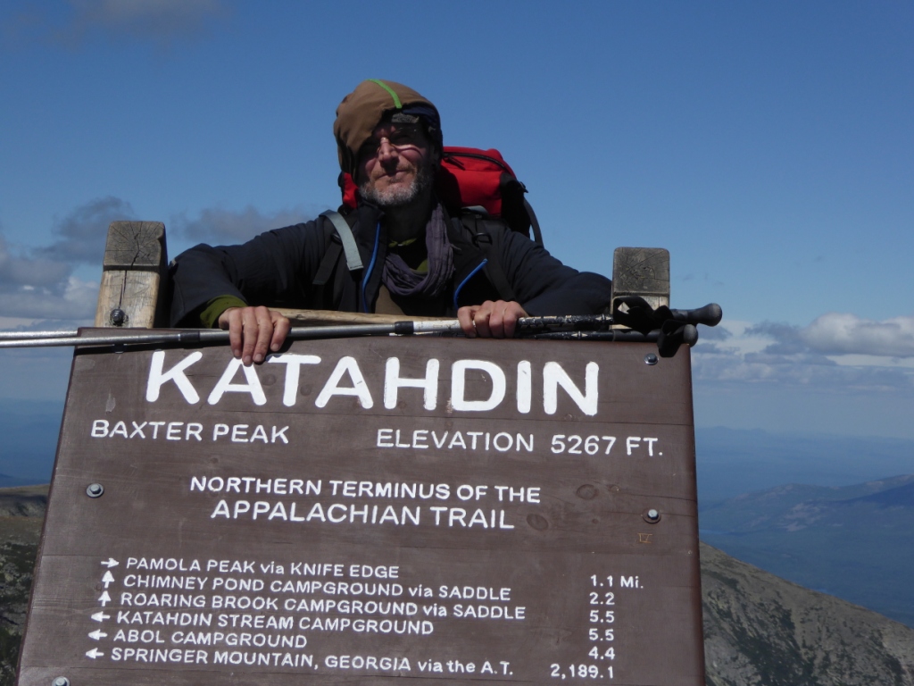 Ein Wanderer lehnt über einem großen Holzschild. Auf dem Schild ist der Gipfel des Katahdin-Berges in Maine angezeigt, die Höhe des Berges. Außerdem steht dort, dass dieser Punkt das nördliche Ende bzw. den nördlichen Start des Appalachian Trails ist. Darunter sind noch einige Kilometerangaben zu weiteren Zielen angegeben. Der Wanderer wirkt müde, er trägt einen großen Rucksack auf dem Rücken.