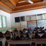 Das Bild zeigt ein Klassenzimmer in Afrika. Die meisten Schüler haben ihren Kopf auf den Tisch gelegt. Im Hintergrund sind zwei große Plakate mit Zahlen und Buchstagen zu sehen. Es handelt sich um eine Vorschule.