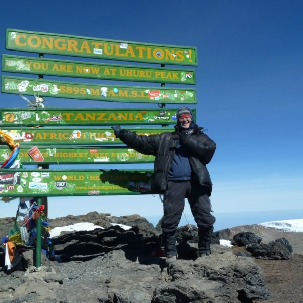 Das Bild zeigt einen sehr warm eingepackten Mann auf dem Gipfel des Kilimanjaro in Tansania. Der Mann zeigt auf verschiedene grüne Schilder, welche den Gipfel kennzeichnen und die Wanderer beglückwünschen. Rechts im unteren Teil des Bildes ist ein Gletscher zu sehen.