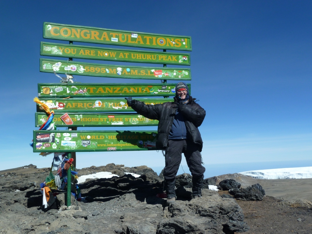 Das Bild zeigt einen sehr warm eingepackten Mann auf dem Gipfel des Kilimanjaro in Tansania. Der Mann zeigt auf verschiedene grüne Schilder, welche den Gipfel kennzeichnen und die Wanderer beglückwünschen. Rechts im unteren Teil des Bildes ist ein Gletscher zu sehen.
