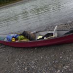 Das Bild zeigt im Vordergrund ein Kanu, welches mit Gepäck beladen ist und am Ufer eines Flusses steht.