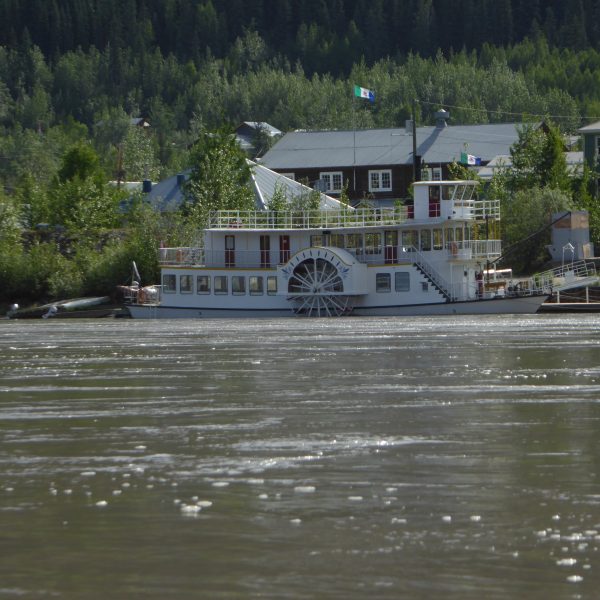 Das Bild zeigt einen Fluss im Vordergrund. Auf dem Fluss ist in der Mitte ein alter Schaufelraddampfer zu sehen. Dahinter sind Häuser zu sehen, dahinter ein Wald. Das Schiff liegt an einem Steg.