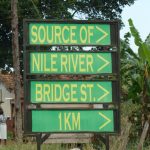 Das Bild zeigt ein Schild, auf dem "Source of Nile River Bridge St. 1 KM" zu lesen ist. Rechts vom Schild sind Bananenstauden zu sehen, links vom Schild Menschen vor Gebäuden.