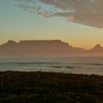 Das Bild zeigt den Tafelberg in Kapstadt während des Sonnenuntergangs, aufgenommen aus einer Entfernung von ungefähr einem Kilometer. Im Bild ist Strand zu sehen und ein Streifen Meer.