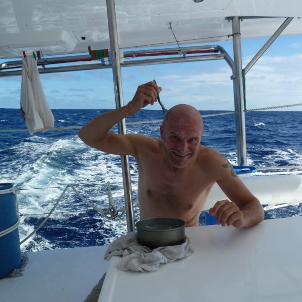 Das Bild zeigt in der Mitte einen Mann mit nacktem Oberkörper am Heck eines Bootes. Der Mann rasiert sich den Kopf mit einem Nassrasierer. Im Hintergrund sind kleine Wellen auf dem Meer zu erkennen, die hinter dem Boot mit Gischt versehen sind. Am Horizont erkennt man auf der linken Seite teils blauen Himmel, während auf der rechten Seite ein zusammenhängendes Band von weißen Wolken zu sehen ist. Auf der linken Seite ist ein blaues Fass auf dem Boot, welches an einem Gestänge befestigt ist. Vor dem Mann steht ein kleiner Stahltopf mit Wasser, der auf einem weißen Lappen abgestellt ist, welcher wohl den weißen Tisch schützen soll.
