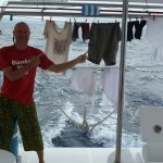 Das Bild sieht das Heck eines Bootes. Im Hintergrund ist das Meer mit leichten Wellen zu sehen. Auf der linken Seite steht ein Mann mit rotem T-Shirt und grüner Short und zeigt lächelnd mit beiden Händen auf Wäsche, die er zwischen Zwei Pfosten an einer Leine zum trocknen aufgehängt hat.