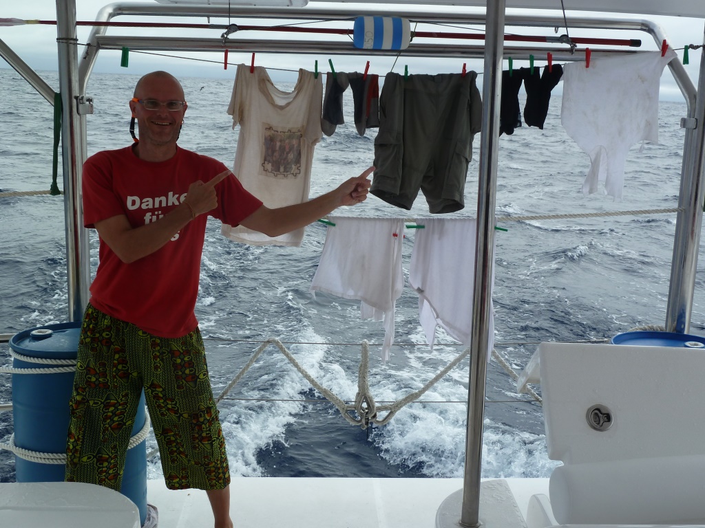 Das Bild sieht das Heck eines Bootes. Im Hintergrund ist das Meer mit leichten Wellen zu sehen. Auf der linken Seite steht ein Mann mit rotem T-Shirt und grüner Short und zeigt lächelnd mit beiden Händen auf Wäsche, die er zwischen Zwei Pfosten an einer Leine zum trocknen aufgehängt hat.