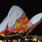 Das Bild zeigt die Oper in Sydney. Im Rahmen des Festival des Lichts wird diese von Beamern farbig angestrahlt, zu sehen sind die unteren Teile in Kunterbunt, während von oben weiße Sahne darüberläuft.