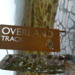 Das Bild liegt auf der linken Seite (gekippt). Es zeigt in der Mitte ein Metallschild, aus dem "Overland-Track" herausgefräst ist. darunter sieht man einen steineren Obelisken, an dem das Schild befestigt ist. Im Hintergrund ist eine Graslandschaft zu sehen, im rechten teil einen Teil eines gepflasterten Weges.