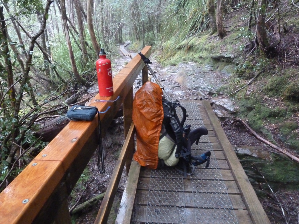 Das Bild zeigt einen hölzernen Steg, der mit einem Handlauf auf der linken Seite von vorne zur Mitte hin mittig durch das Bild läuft. Auf dem Steg steht ein großer Rucksack in einer Regenhülle, dahinter sind zwei Wanderstöcke an den Handlauf gelehnt, auf dem auch die blaue Hülle eines Fotoapparates, eine Brille sowie eine rote Wasserflasche liegen. Der Weg im Hintergrund zeigt Spuren von Schnee auf, rechts und links des Weges sind Bäume zu sehen. Der Steg ist mit Hühnerdraht versehen, vermutlich um ein Ausrutschen der Wanderer zu verhindern.