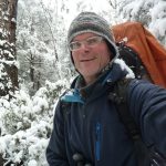 Das Bild zeigt einen Mann, warm eingepackt mit Mütze und einem großen Rucksack in einem verschneiten Wald. Schnee liegt auch auf dem Kragen seiner Wanderjacke. Der Mann lächelt in die Kamera.