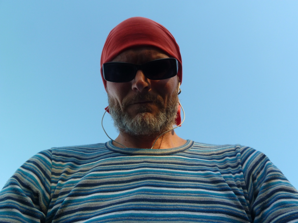 Das Bild zeigt einen Mann in einem geringelten Pullover und mit einer roten Kopfbedeckung. Der Mann trägt eine schwarze Sonnenbrille und hat Kopfhörer in den Ohren. Im Hintergrund ist lediglich blauer Himmel zu sehen.