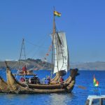 Das Bild zeigt ein großes Schilfboot auf dem Titicaca-See in Bolivien. Das Boot besteht aus zwei bananenförmig gebogenen Schilfrümpfen, zwischen denen eine Plattform befestigt ist. Im vorderen Bereich des Bootes ist ein Segelmast, dessen Segel gehisst ist. Am oberen Ende des Mastes weht die Flagge Boliviens. Ebenfalls vorne ist ein Mann mit einem großen Ruder zu sehen. Im Hintergrund sind Berge mit schneebedeckten Gipfeln zu sehen. Auf dem Boot befinden sich mehrere Menschen. Hervorstechend ist das klare blaue Wasser des Sees im unteren Drittel des Bildes.