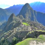 Das Bild zeigt die alte Inka-Stadt Macchu Picchu von einer erhöhten Warte aus. Mittig im Bild sind die Ruinen der Stadt zu erkennen, hinter den Ruinen erhebt sich der Huayna Picchu-Berg, der über den Ruinen thront. Im Hintergrund sind weitere, bewaldete Berge zu sehen. In der Mitte der Ruinen ist eine große grüne Grasfläche zu sehen, auch zur rechten Seite sind grüne Terrassen zu sehen.