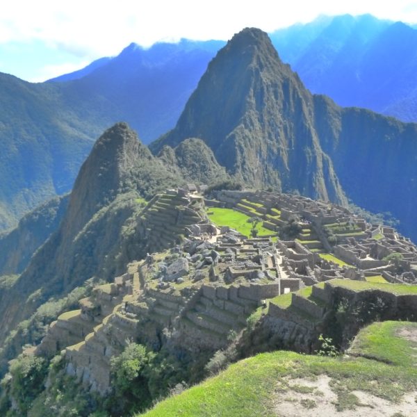 Das Bild zeigt die alte Inka-Stadt Macchu Picchu von einer erhöhten Warte aus. Mittig im Bild sind die Ruinen der Stadt zu erkennen, hinter den Ruinen erhebt sich der Huayna Picchu-Berg, der über den Ruinen thront. Im Hintergrund sind weitere, bewaldete Berge zu sehen. In der Mitte der Ruinen ist eine große grüne Grasfläche zu sehen, auch zur rechten Seite sind grüne Terrassen zu sehen.