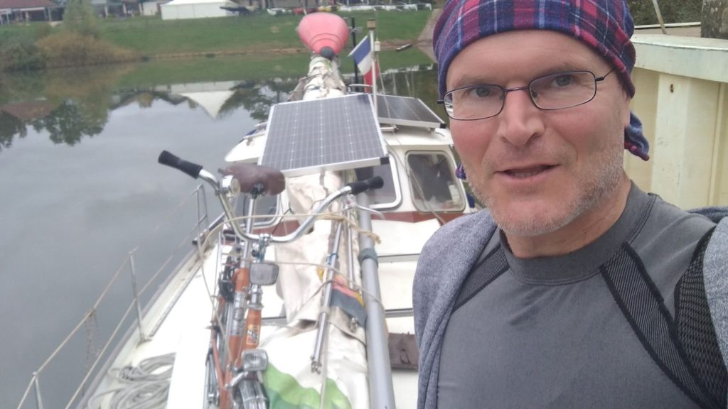 Das Bild zeigt einen Mann auf einem Segelboot. Auf dem Segelboot ist ein Fahrrad befestigt. Auch zwei größere Solar-Paneele sind auf dem Boot befestigt.