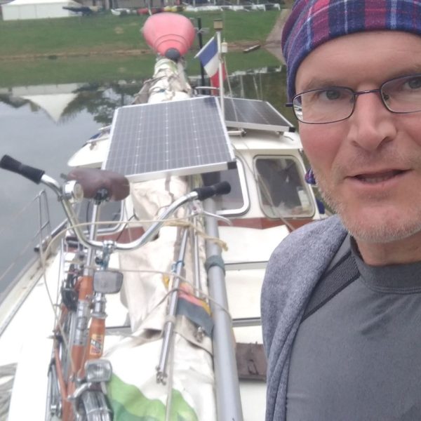 Das Bild zeigt einen Mann auf einem Segelboot. Auf dem Segelboot ist ein Fahrrad befestigt. Auch zwei größere Solar-Paneele sind auf dem Boot befestigt.
