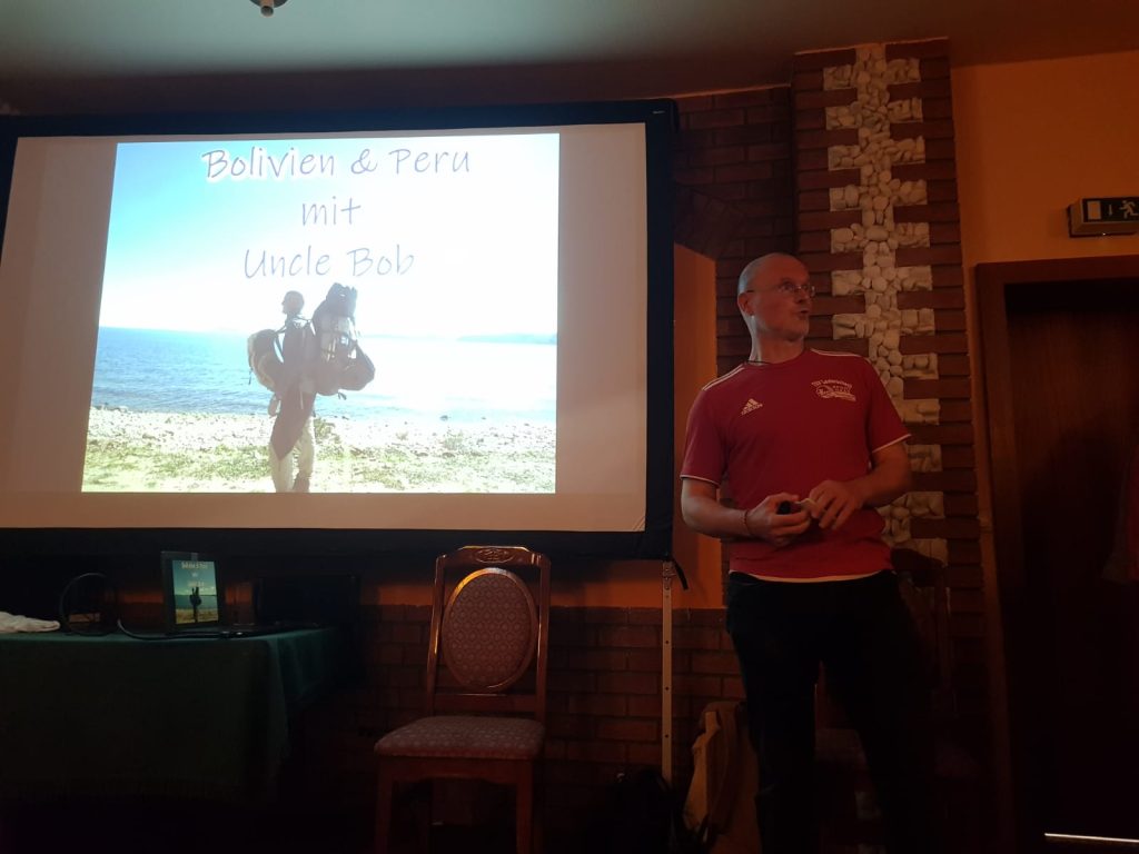 Das Bild zeigt einen Mann, der in einem Raum vor einer Leinwand steht. Auf diese Leinwand ist ein Bild gestrahlt, dass diesen Mann mit Rucksäcken an einem See zeigt. Darüber ist der Titel "Bolivien & Peru mit Uncle Bob" abgebildet. Vor der Leinwand steht ein Stuhl.