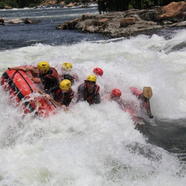 Das Bild zeigt ein Rafting-Boot mit Menschen darin, wie es in einem Fluss in eine Stromschnelle gerät und von dieser überspült wird. im Hintergrund sind Felsen am Ufer und zum Teil im Wasser zu sehen.