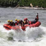 Das Bild zeigt ein Rafting-Boot, welches auf einer Stromschnelle reitet. Während sich die Gäste in dem Boot alle an die Rettlungsleine klammern, winkt der Steuermann lustig mit dem hochgestreckten Ruden in die Kamera. Links vom Boot ist ein umgekipptes Rafting-Boot zu sehen, dahinter eine Mann in einem kleinen Kajak, dahinter ein weiteres Rafting-Boot. Im oberen Teil des Bildes sieht mal felsiges Ufer, dahinter Büsche.