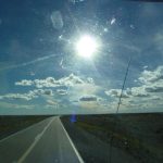 Das Bild zeigt eine Straße, die durch eine karge Landschaft führt. Es wurde durch eine Autoscheibe aufgenommen, teilweise sind schlieren und ein Steinschlag zu erkennen. Zwei Drittel des Bildes nimmt der Himmel ein, der von ein paar weißen Wolken bedeckt ist.