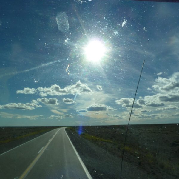 Das Bild zeigt eine Straße, die durch eine karge Landschaft führt. Es wurde durch eine Autoscheibe aufgenommen, teilweise sind schlieren und ein Steinschlag zu erkennen. Zwei Drittel des Bildes nimmt der Himmel ein, der von ein paar weißen Wolken bedeckt ist.