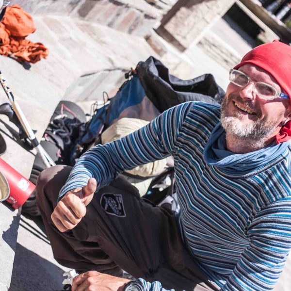 Das Bild zeigt einen Mann mit Bart, geringeltem Pullover und roter Kopfbedeckung. Er zeigt seinen rechten Daumen als Zeichen der Zustimmung. Im linken Bild steht ein Topf mit Essen auf einem Campingkocher. Neben dem Kocher liegen zwei Wanderstöcke. Hinter dem Mann ist ein großer Rucksack zu sehen.